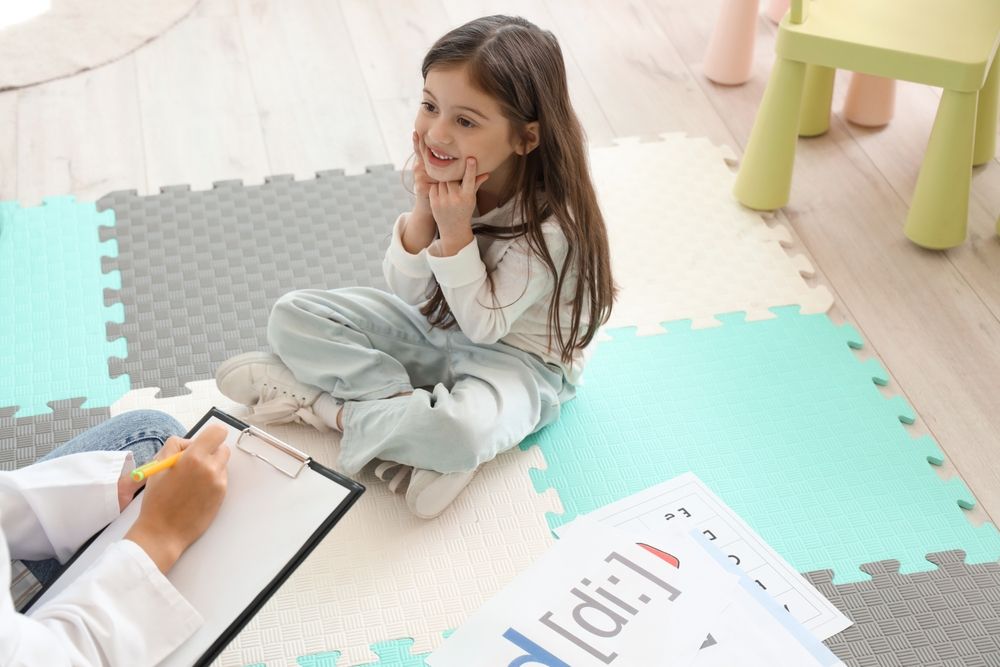 A speech therapist takes session notes while a young girl smiles at her on a colorful puzzle-mat floor